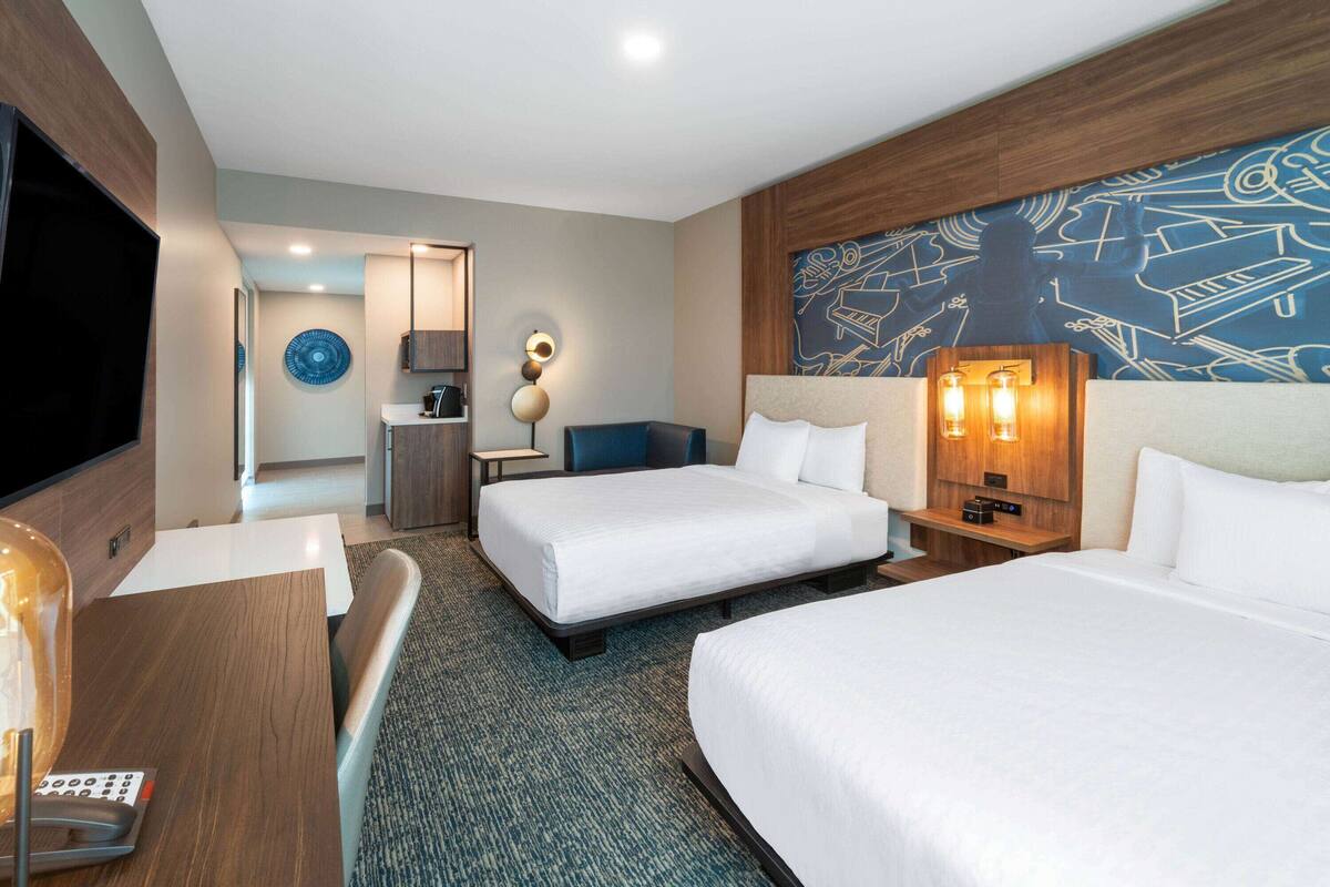 A hotel room with two beds, a desk, a TV, a lamp, and a wall with musical notes.