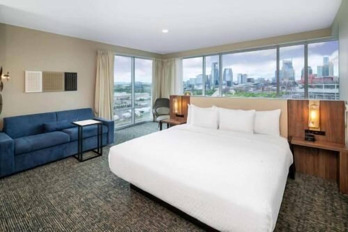 A hotel room with a large bed, a blue sofa, a small table, and a view of a cityscape through large windows.