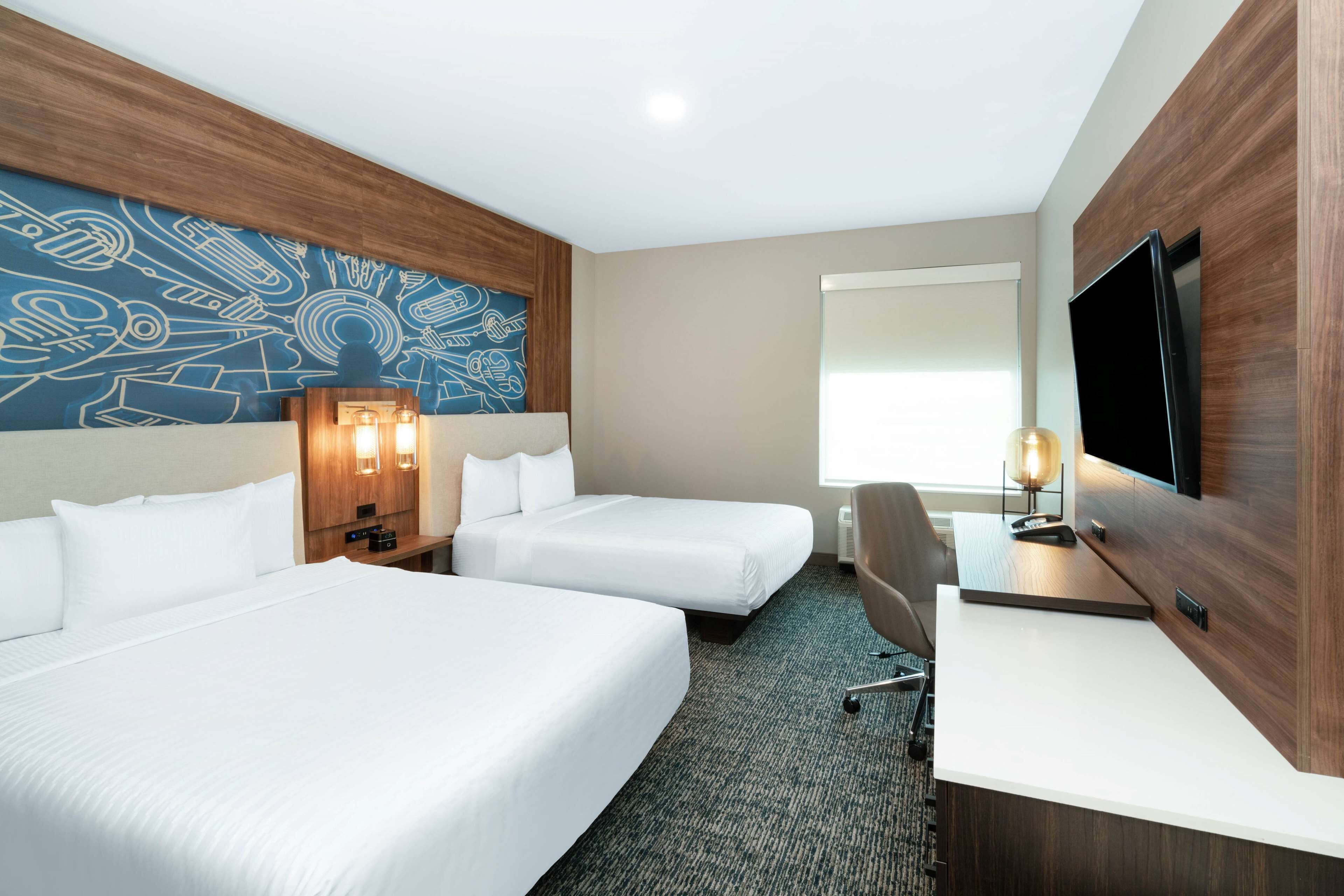 Habitación de hotel con dos camas, un escritorio, un televisor y una pared con un diseño abstracto azul.