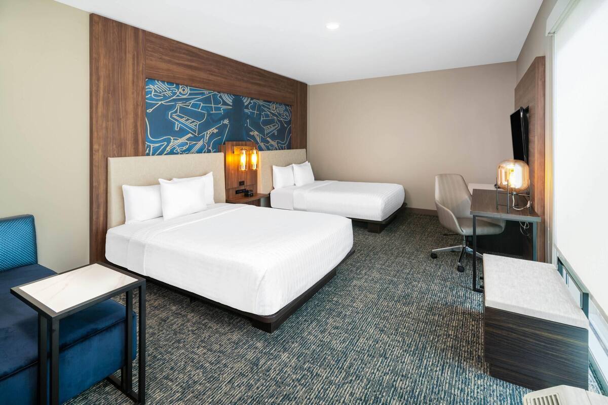 A hotel room with two beds, a desk, a chair, a small table, and a wall-mounted TV.