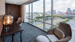 A modern hotel room with a large window offering a city view, a wooden wardrobe, a glass table with a light, and a comfortable armchair.