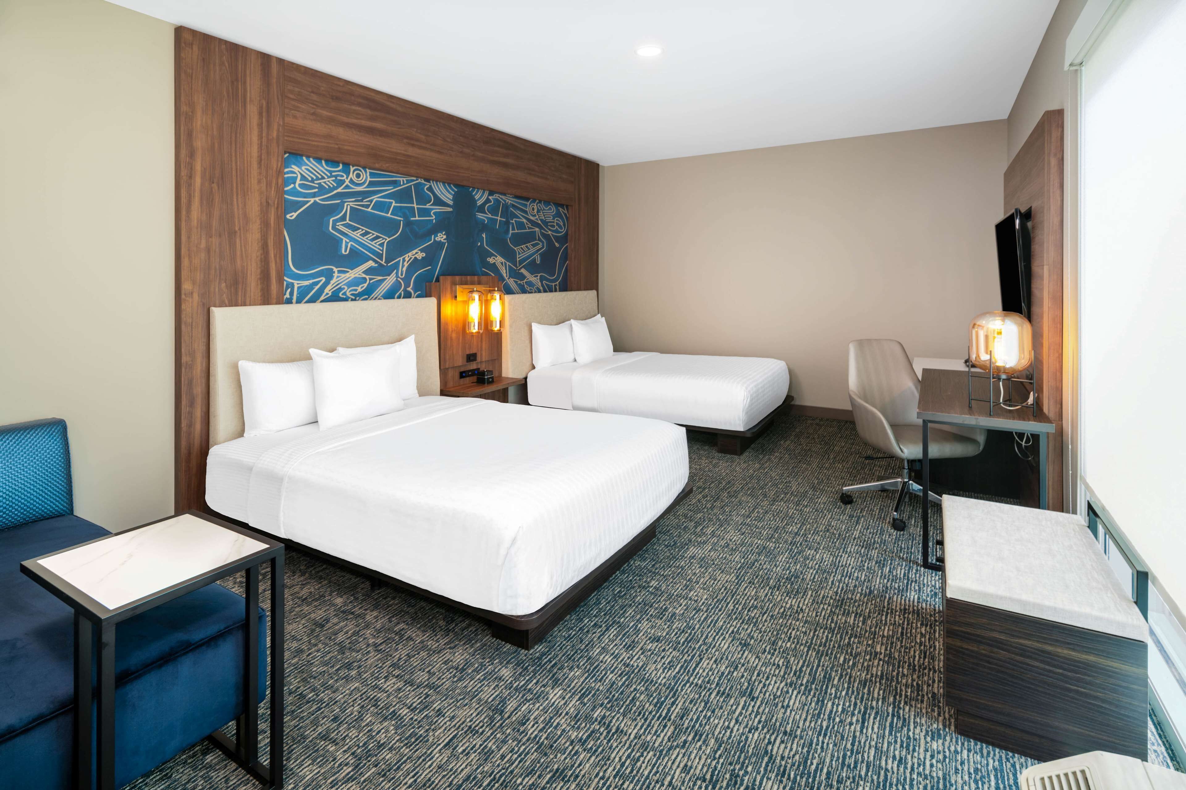 Une chambre d’hôtel avec deux lits, un bureau, une chaise, une petite table et un grand tableau abstrait accroché au mur.