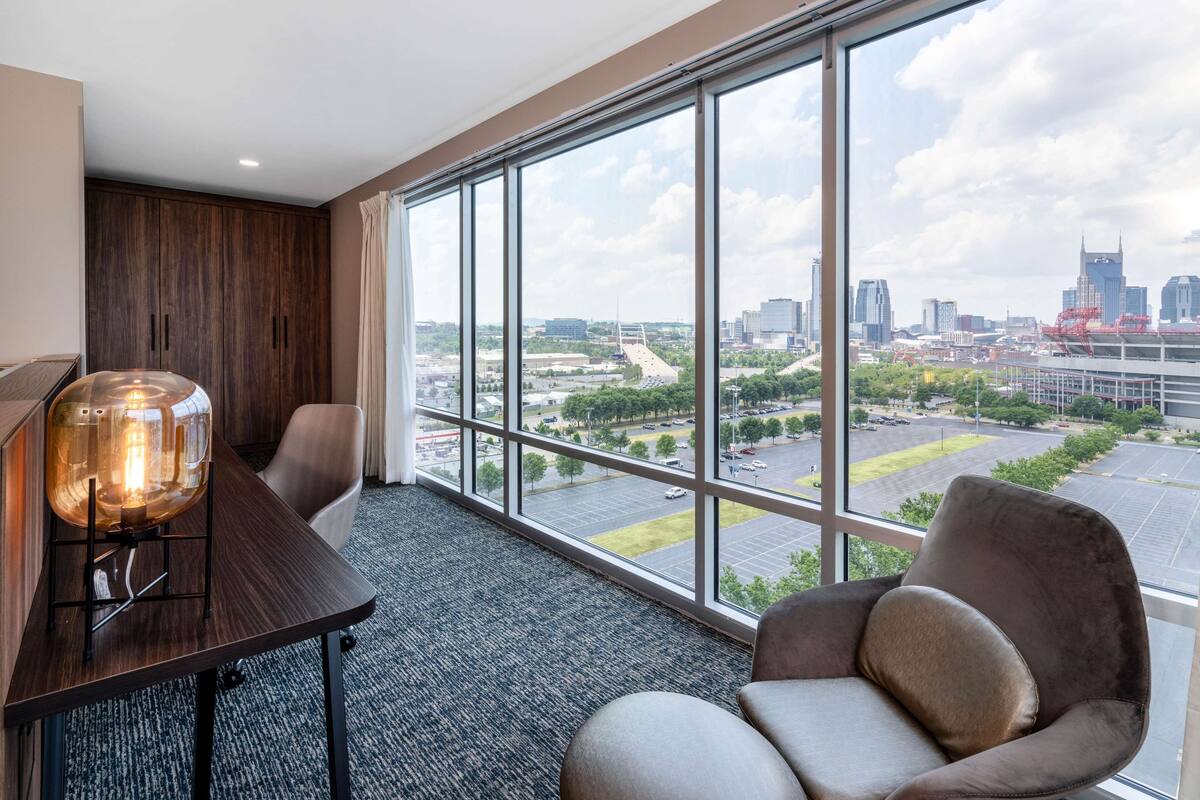 A modern hotel room with a large window, a wooden wardrobe, a table with a glass lamp, and a comfortable chair.