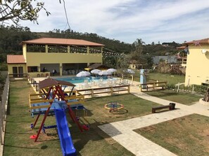 A playground with a slide and swings, a pool area with umbrellas, and a building with a tiled roof. - Site with chalets and wet bar (Igaratá)