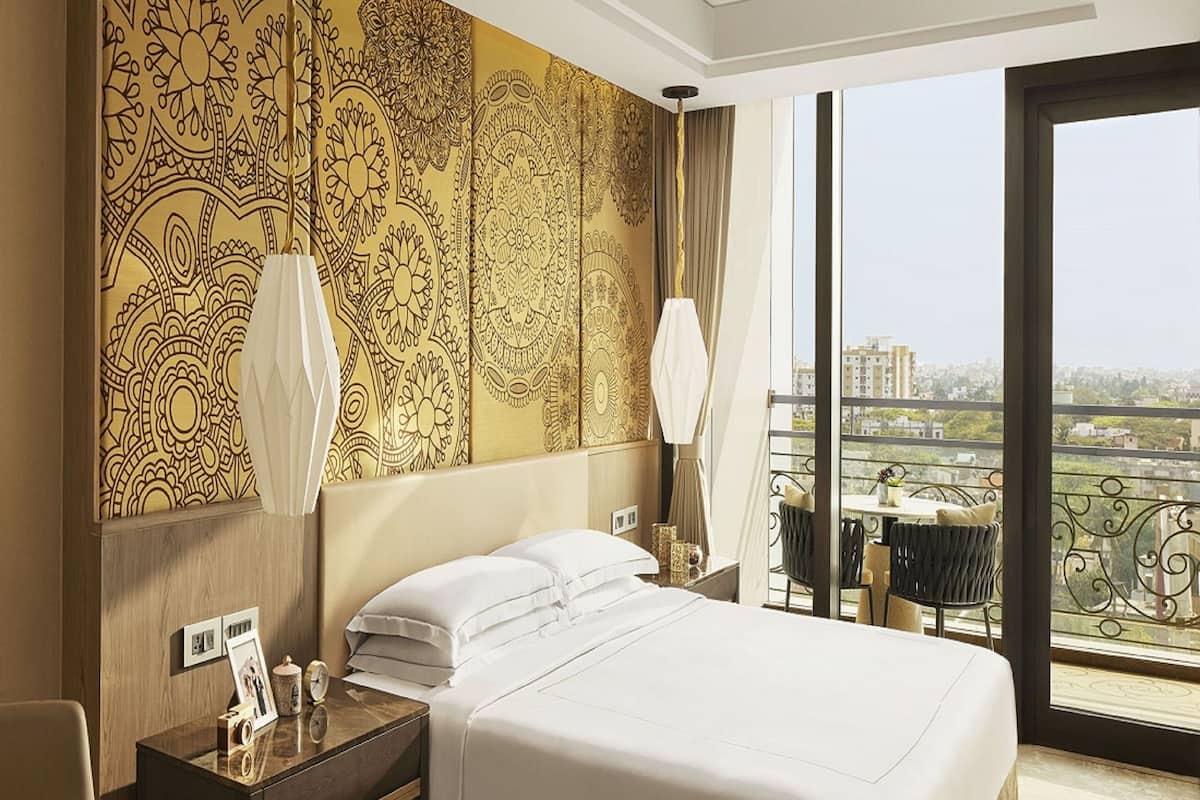Une chambre d’hôtel avec un grand lit, une table de chevet avec une lampe, une vue sur la ville grâce à de grandes fenêtres et un mur décoré d’un motif.