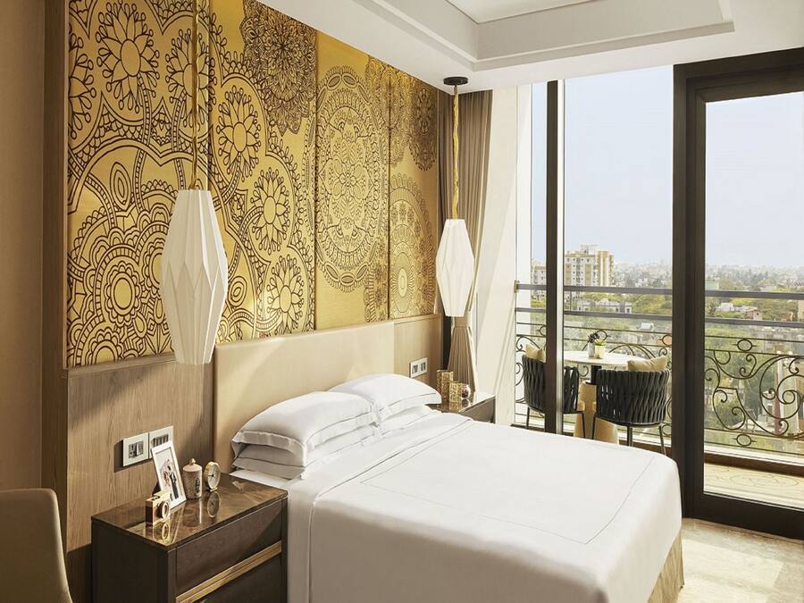 Una habitación de hotel con una cama grande, una mesita de noche con lámpara, vistas de la ciudad a través de amplios ventanales y un papel tapiz estampado.