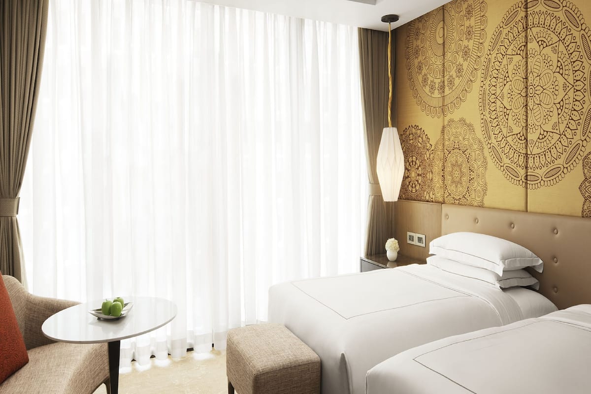 Une chambre d’hôtel avec deux lits, une petite table sur laquelle est posé un vase, et une grande fenêtre avec des rideaux légers.