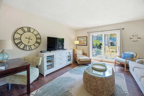 A living room with a large wall clock, a flat-screen TV, wicker furniture, a glass coffee table, and a view of a building labeled 'BEACH HOUSE'.