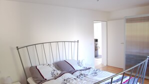 A single bed with a metal frame, white bedding, and a pillow with a leaf pattern. There is a bedside table with a lamp and a power outlet. The room has white walls and a wooden floor. - Wohnung mit Kamin Terrasse und Grill in der Nähe von Nürnberg (Burgthann)