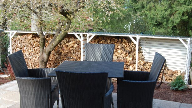 An outdoor patio with a table and four chairs, a tree, and a stack of firewood. - Wohnung mit Kamin Terrasse und Grill in der Nähe von Nürnberg (Burgthann)