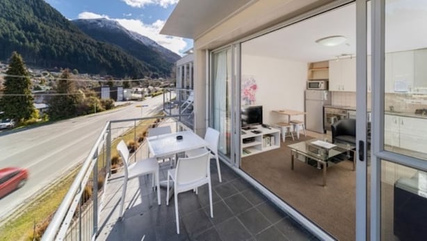 A modern apartment with a balcony overlooking a road and mountains. It includes a dining area, kitchen, and a living space with a TV and books. - Mountain View City Aparment (Queenstown)