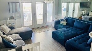 A spacious living room with a blue sofa set, a white chair, and a ceiling fan. - Family Time Oceanfront Condo at Ocean Isle (Ocean Isle Beach)