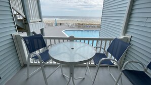 A balcony with a glass table and chairs, overlooking a swimming pool and beach. - Family Time Oceanfront Condo at Ocean Isle (Ocean Isle Beach)
