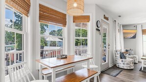 Un coin repas avec une table en bois et des chaises blanches, de grandes fenêtres équipées de stores et un luminaire suspendu en osier.