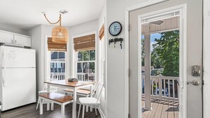 Une cuisine avec un réfrigérateur blanc, une table à manger en bois et des chaises, un luminaire suspendu et une porte donnant sur une terrasse extérieure.