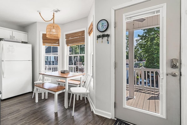 A kitchen with a white refrigerator, wooden dining table, and chairs, a hanging light fixture, and a door leading to an outdoor deck.