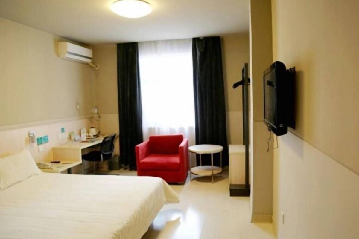 A hotel room with a bed, a desk with a chair, a red armchair, a small round table, and a TV mounted on the wall.