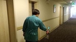 Une personne vêtue d’une veste turquoise marche dans un couloir aux murs de couleur beige et au sol recouvert de moquette, et elle tient un flacon pulvérisateur à la main.