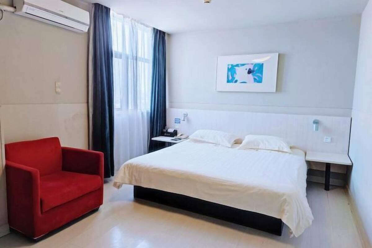 A hotel room with a bed, a red armchair, a nightstand, and a wall-mounted artwork.