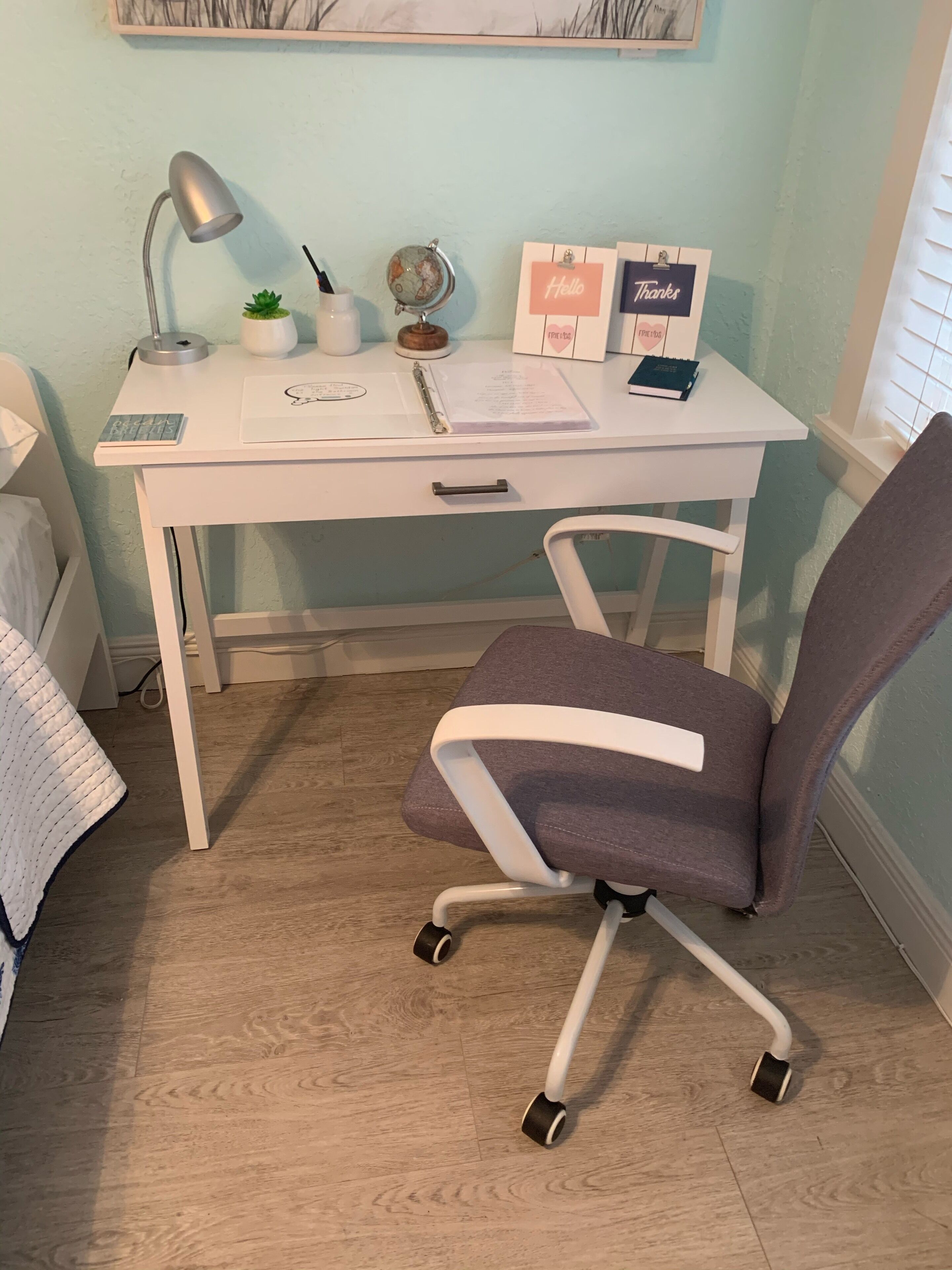 Un bureau blanc avec une chaise de bureau grise, une lampe, un globe terrestre, une petite plante et quelques cartes de remerciement.
