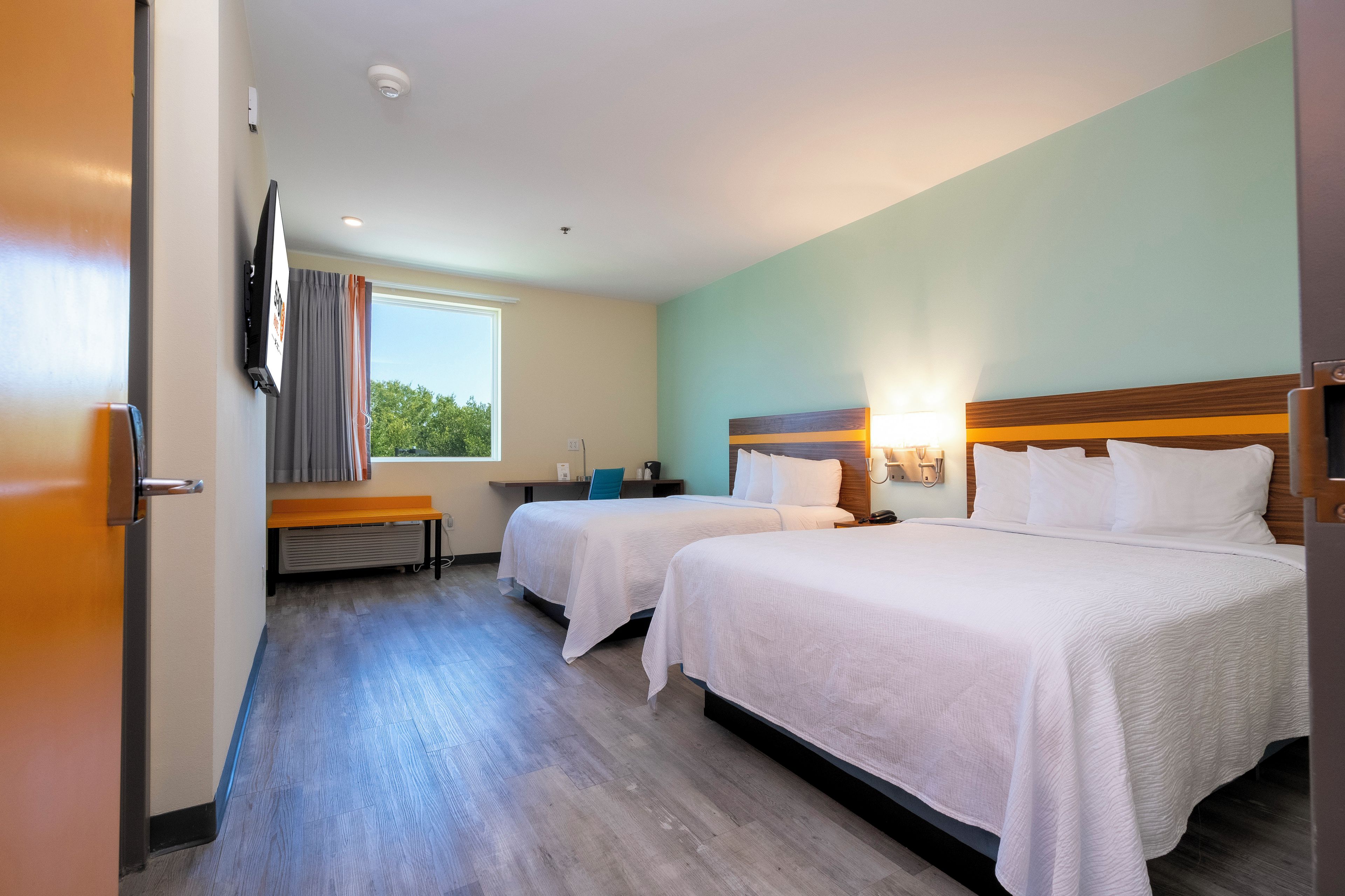 Quarto de hotel com duas camas, uma escrivaninha, uma cadeira, janela com cortinas e TV de tela plana na parede.