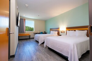 A hotel room with two beds, a desk, a chair, a window with curtains, and a flat-screen TV mounted on the wall.