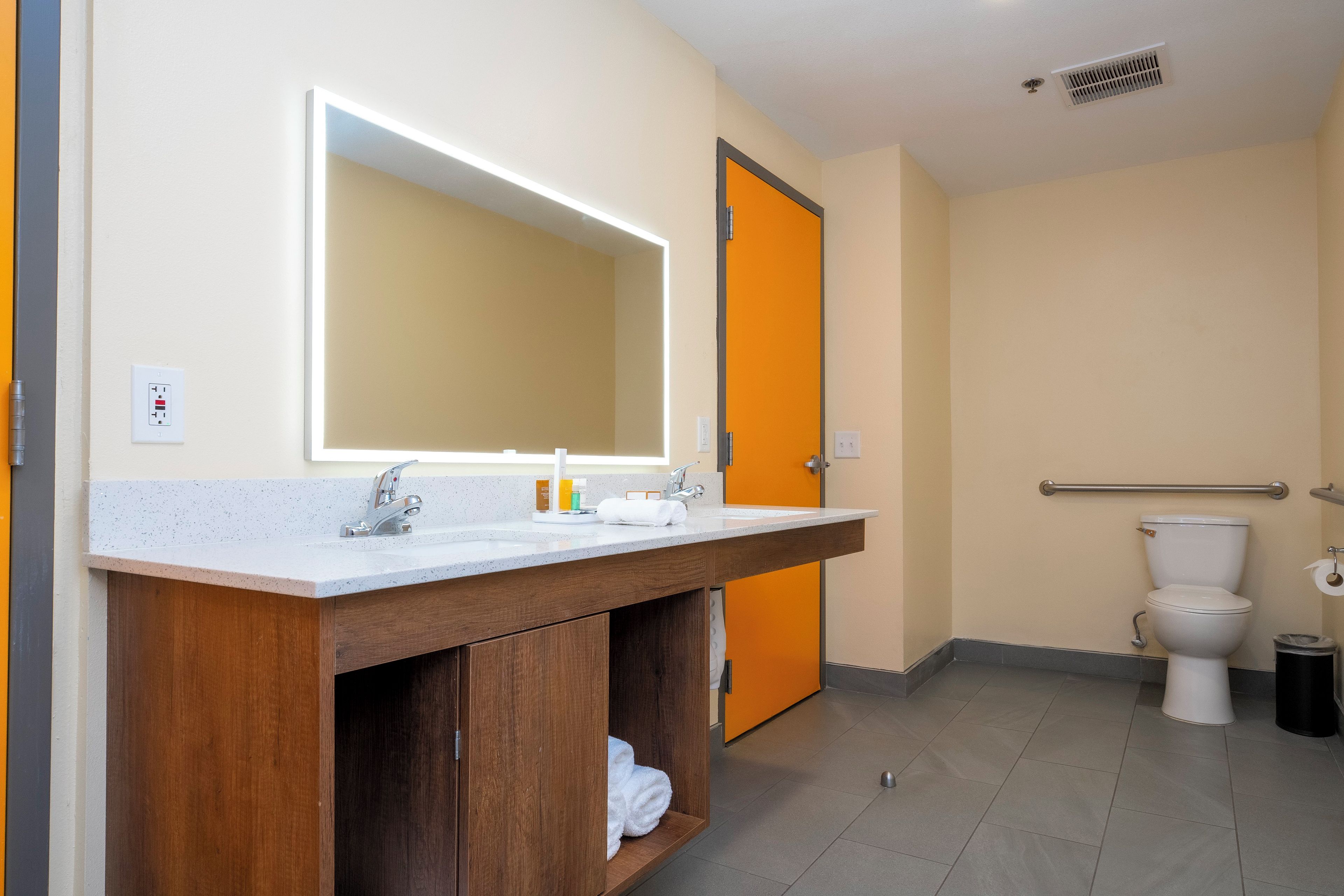 A bathroom with a wooden vanity, a large mirror, a toilet, and a handrail.