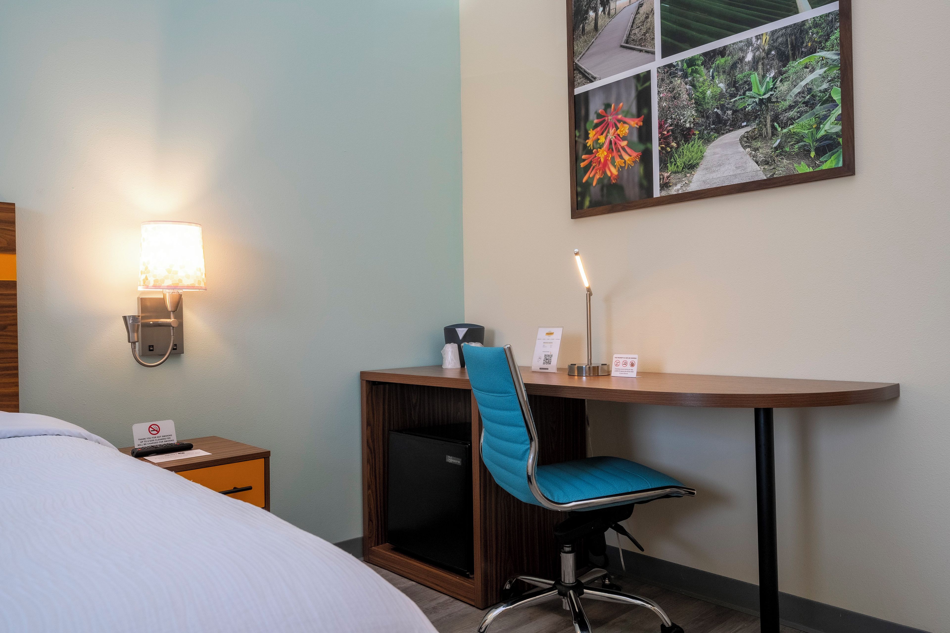 Een hotelkamer met een bed, een bureau met stoel, een wandlamp en een lijst met afbeelding.