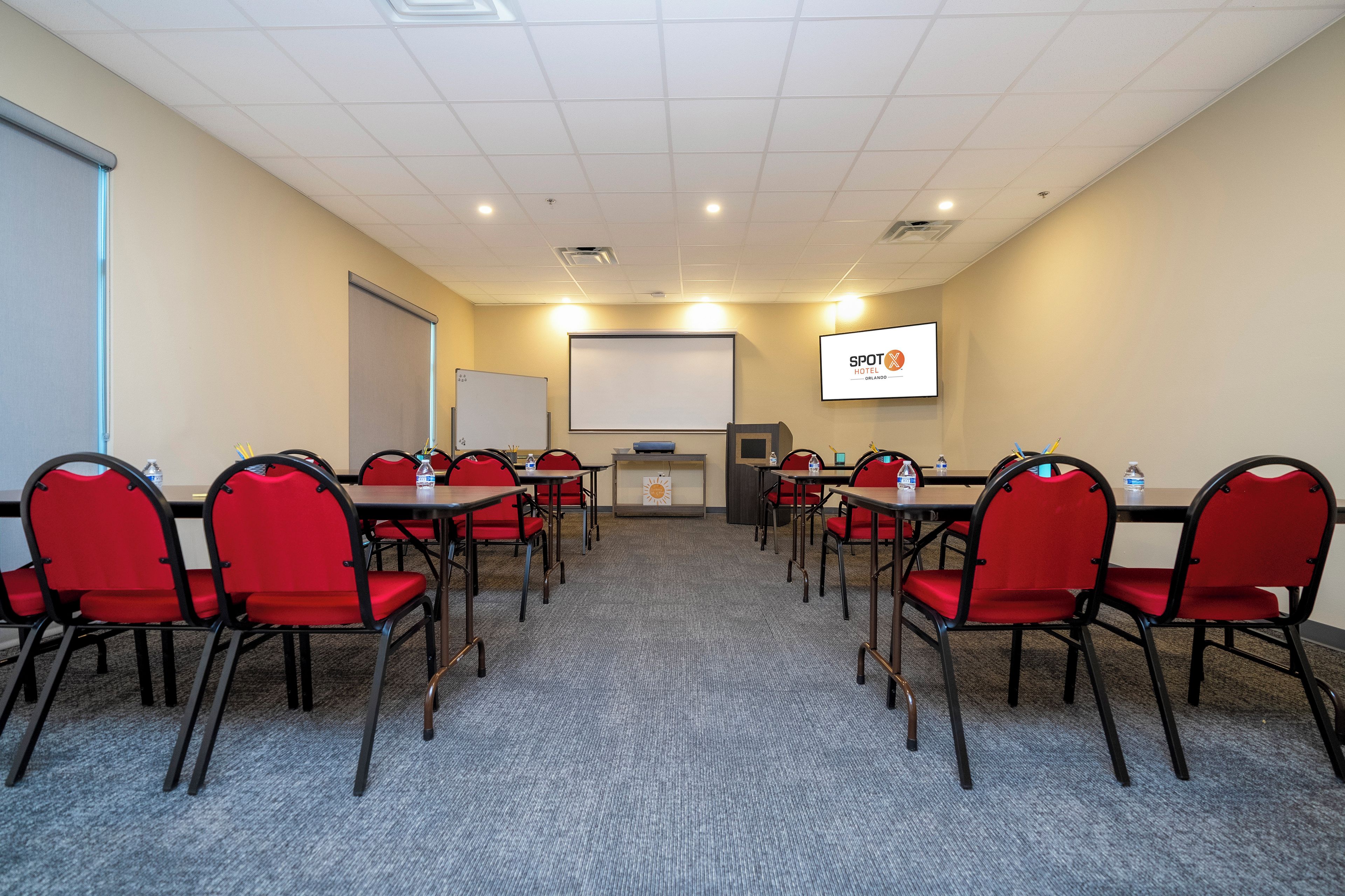 Sala de conferências com cadeiras vermelhas, lousa, tela de projetor e uma garrafa de água sobre a mesa.