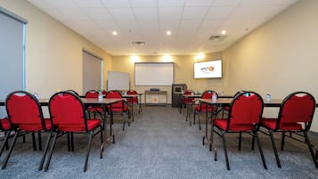 Sala de conferências com cadeiras vermelhas, lousa, tela de projetor e uma garrafa de água sobre a mesa.