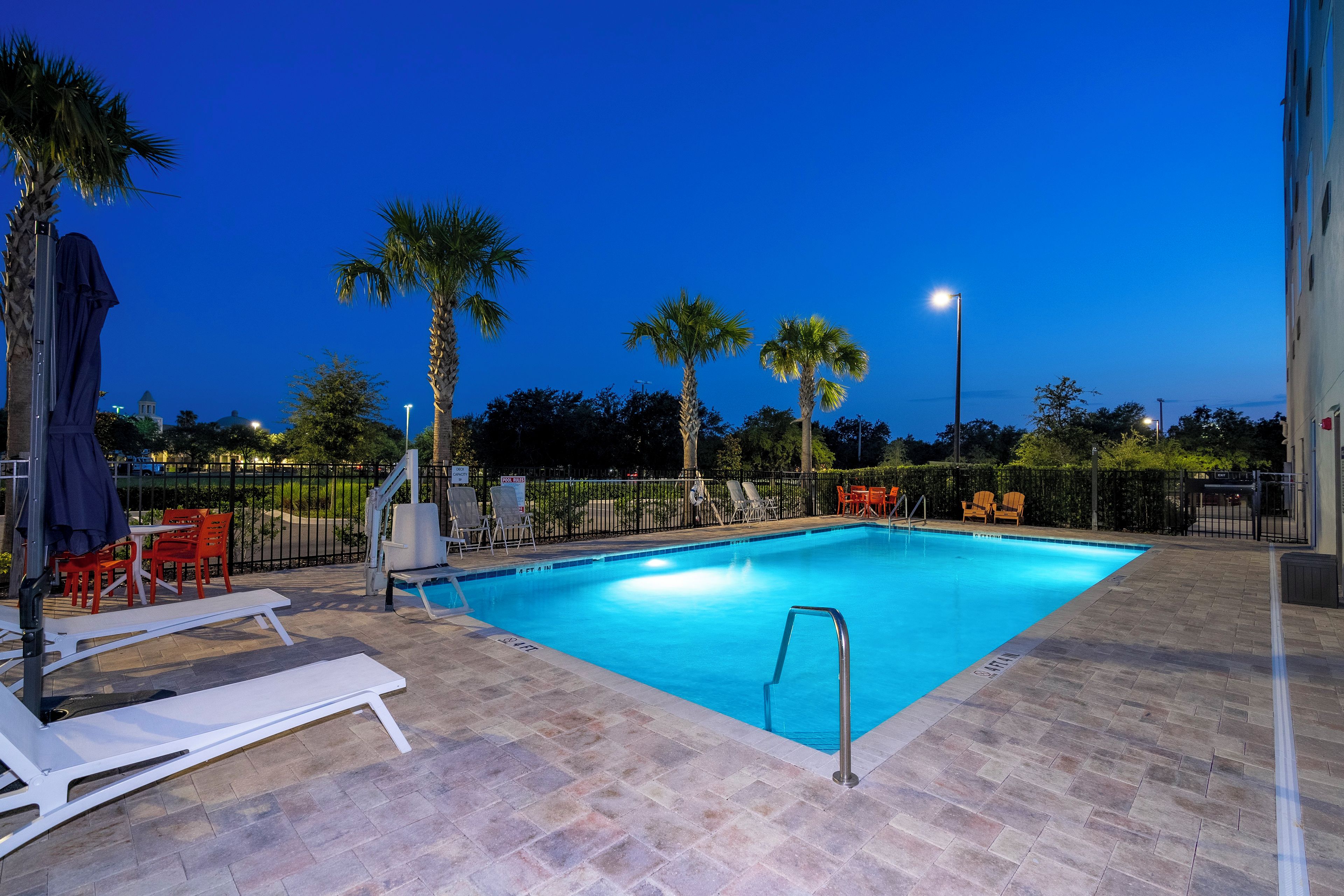 A swimming pool with lounge chairs and a covered seating area, surrounded by palm trees and a fence.