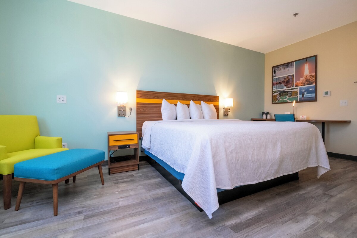 A hotel room with a bed, a yellow armchair, a blue bench, a wooden nightstand, and a wall-mounted lamp.