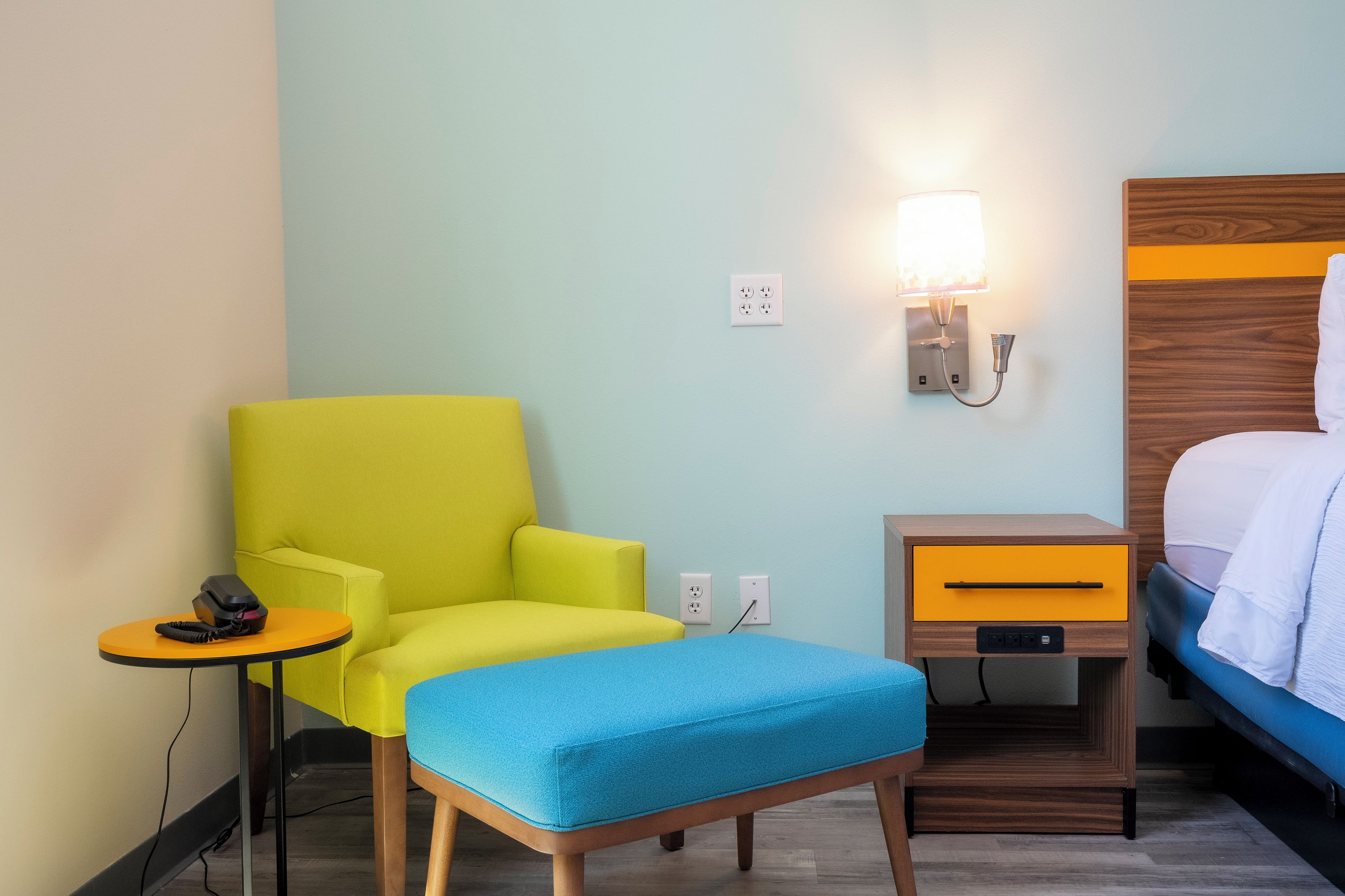 A hotel room with a yellow armchair, a small bedside table with a lamp, a wooden nightstand, and a bed with a blue blanket.
