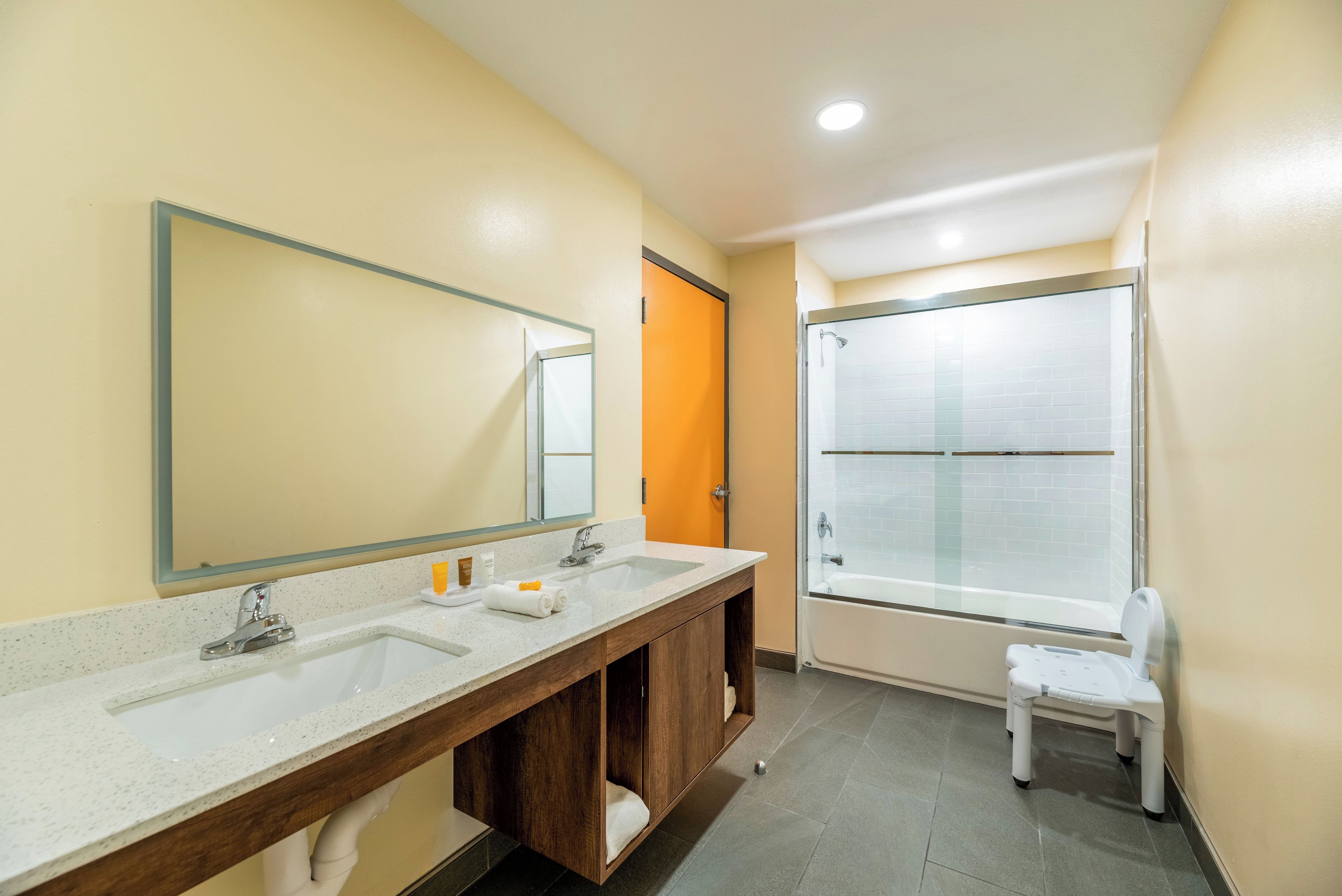 Un baño con lavamanos doble, un espejo grande, una ducha y un taburete pequeño.