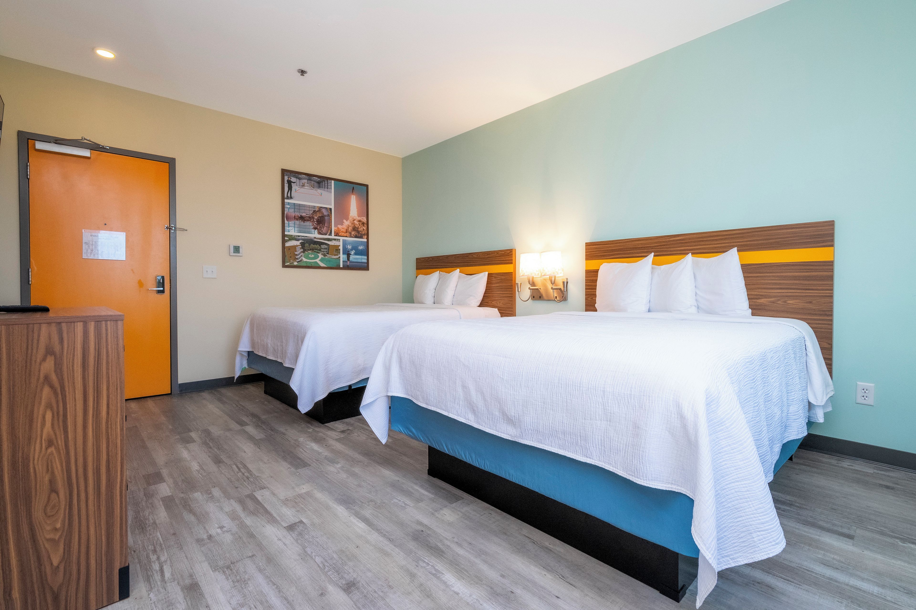 A hotel room with two beds, a wooden headboard, a wall-mounted light fixture, and a door with a sign.