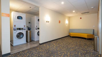 A hotel laundry room with stacked washers and dryers, a bench, and a trash can.