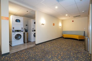 A hotel laundry room with stacked washers and dryers, a bench, and a trash can. - SPOT X Hotel Orlando/Intl Dr by The Red Collection (Orlando)