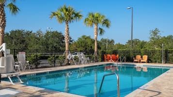 Piscina com escada de metal, cercada por cadeiras de descanso e uma grade.