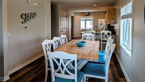 A dining area with a wooden table and white chairs, a kitchen in the background, and a 'Shore' sign on the wall. - Oceanfront 4BR Duplex in Bethany Beach – Panoramic Views & Private Beach Access (Fenwick Island)