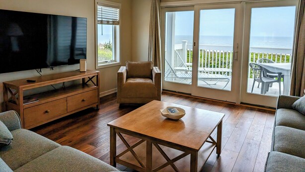 A living room with a TV, a sofa, a coffee table, and a view of the ocean through large sliding doors. - Oceanfront 4BR Duplex in Bethany Beach – Panoramic Views & Private Beach Access (Fenwick Island)