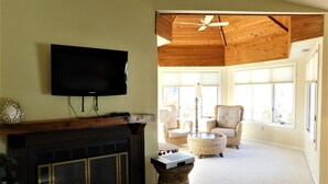 A living room with a fireplace, a flat-screen TV, wicker furniture, and large windows. - Spacious 6BR Family Home – Just 3 Blocks to Bethany Beach in The Canal Community (Bethany Beach)