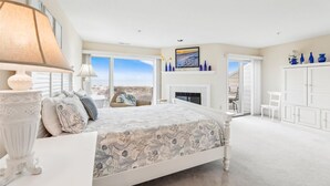Quarto com cama grande, lareira, vista para a praia e um guarda-roupa branco.
