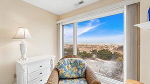 Quarto com uma grande janela que oferece vista para uma praia e dunas, um penteúdo branco, uma cadeira de vime com estofamento floral e uma luminária.