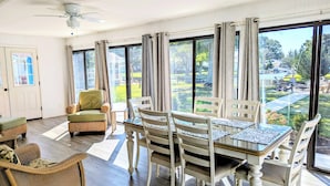 Un comedor con mesa y sillas, un ventilador de techo y ventanas grandes con cortinas.