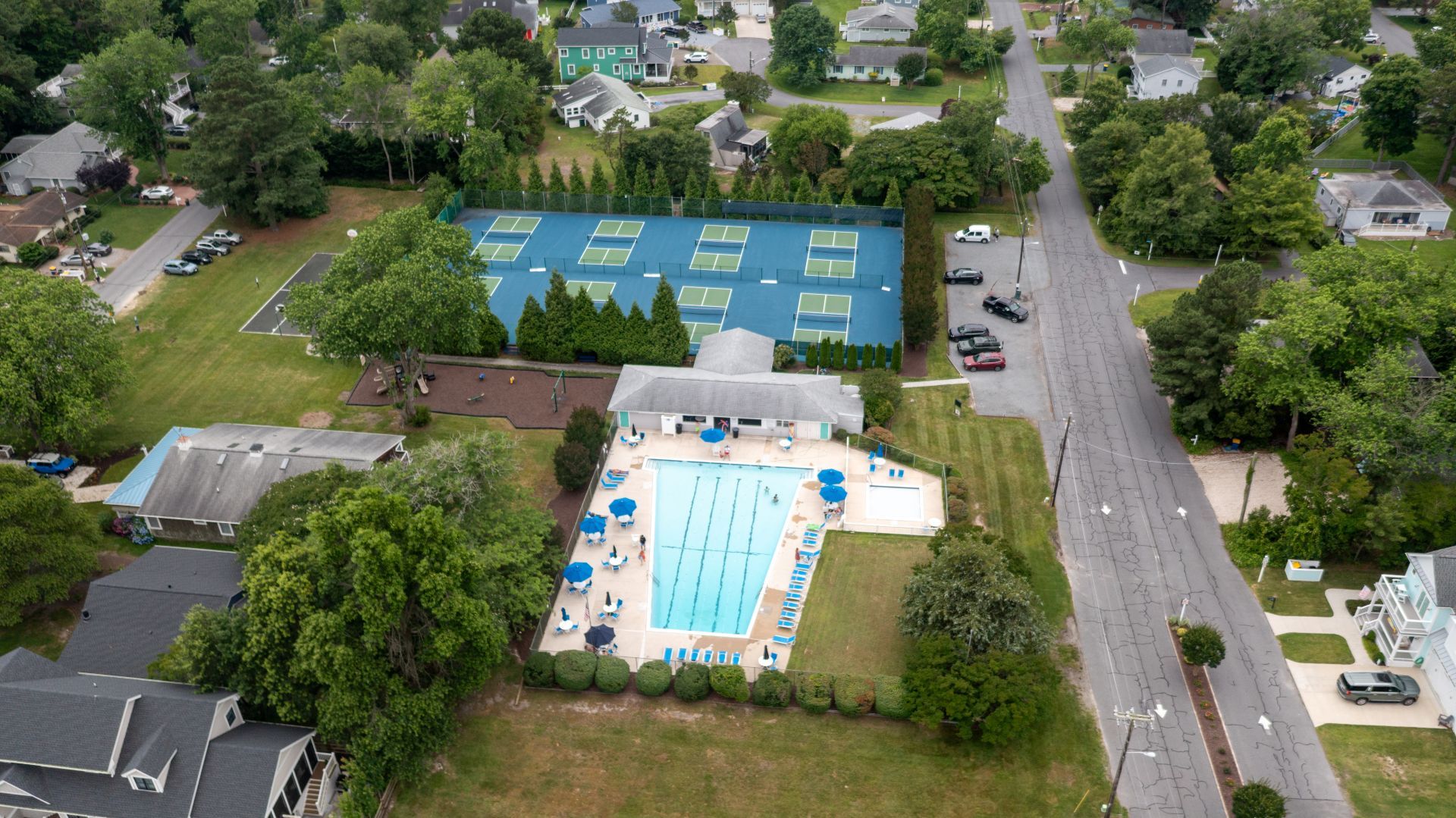 An aerial view of a residential area with a large swimming pool surrounded by a fenced tennis court, a covered pavilion, and a road.