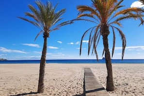 Ein hölzerner Weg führt zu einem Strand mit zwei Palmen und blauem Himmel.