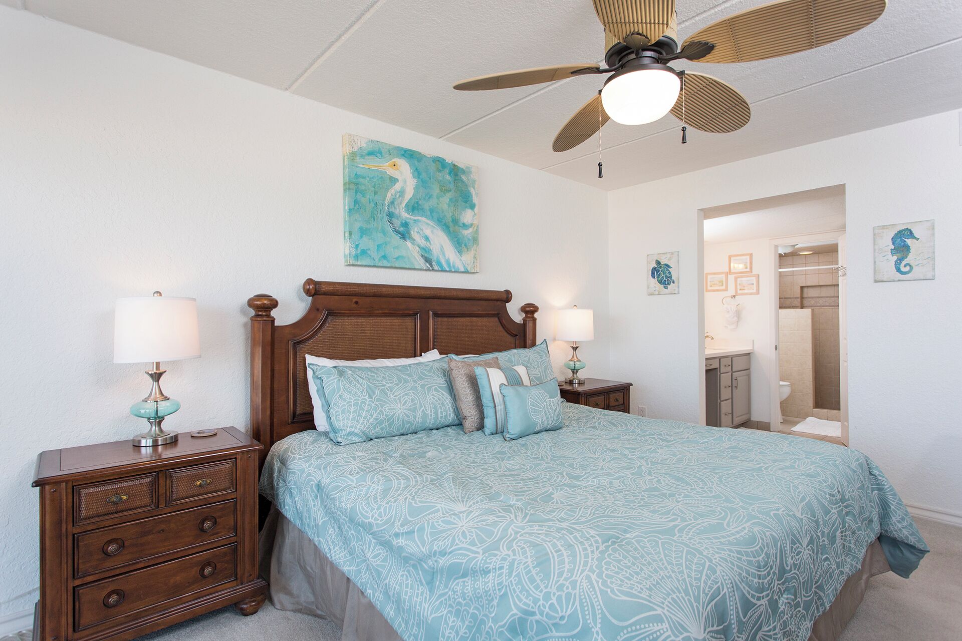 A bedroom with a wooden bed, two bedside tables with lamps, a ceiling fan, and a bathroom visible through an open door.