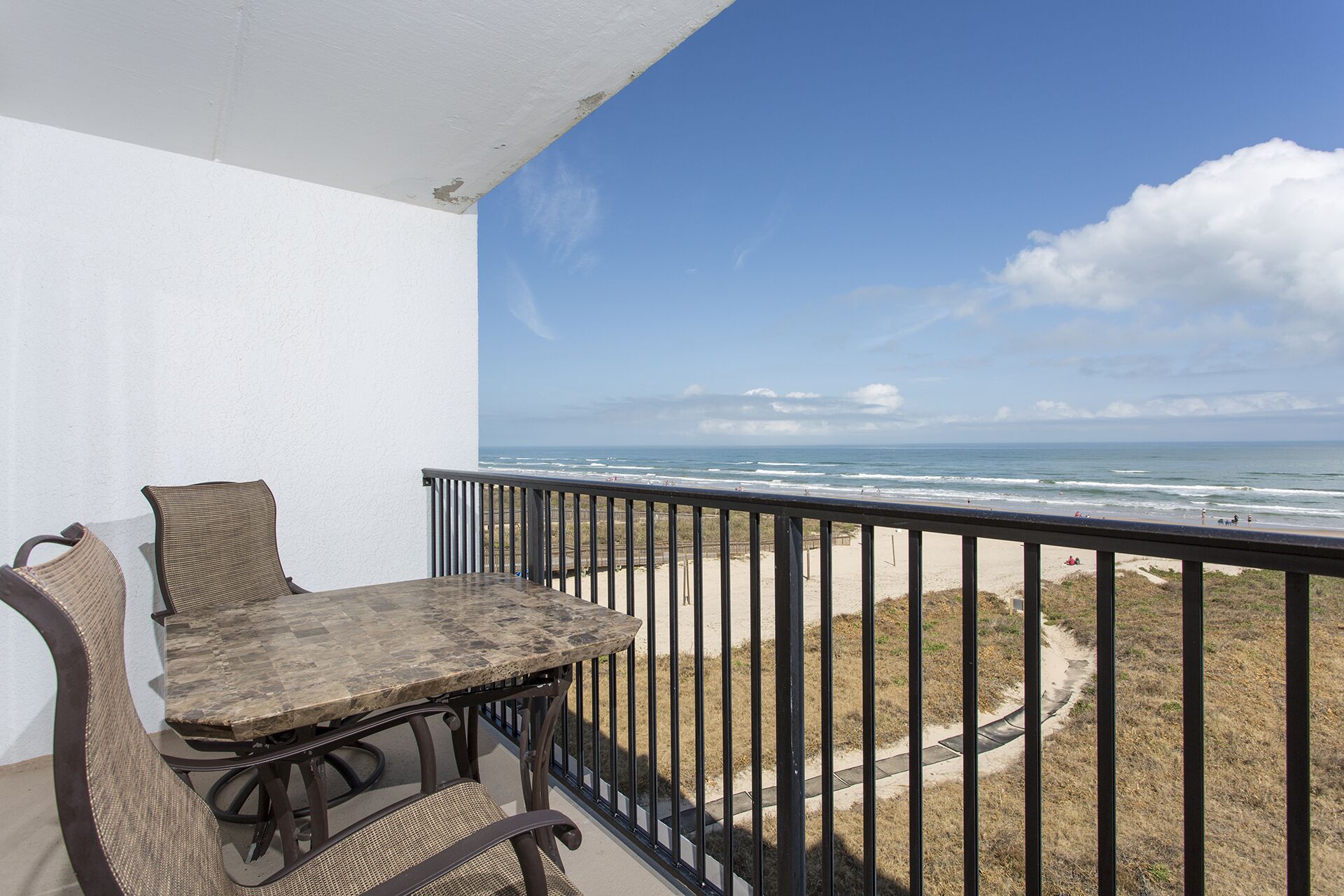 Een balkon met een tafel en stoelen, met uitzicht op een strand met golven en een heldere lucht.