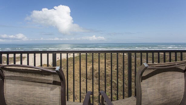 Un balcon avec des fauteuils en osier donnant sur une plage, avec un ciel dégagé et une vue sur l’océan.