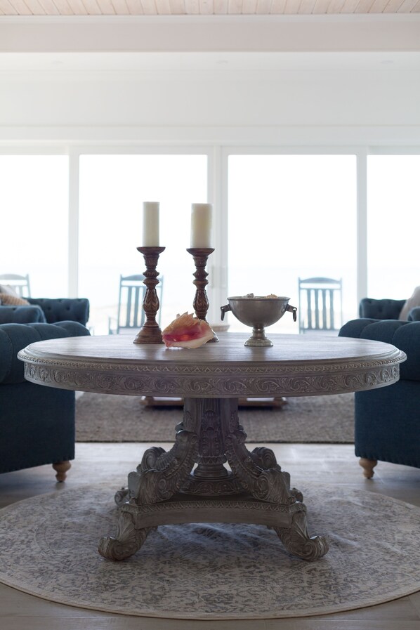 A round table with ornate legs, two candles, and a bowl on a patterned rug in a room with blue upholstered chairs and large windows.